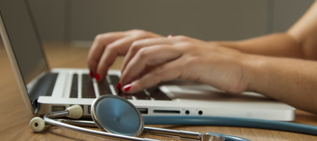 A healthcare administrator works on their laptop with a stethoscope beside them.