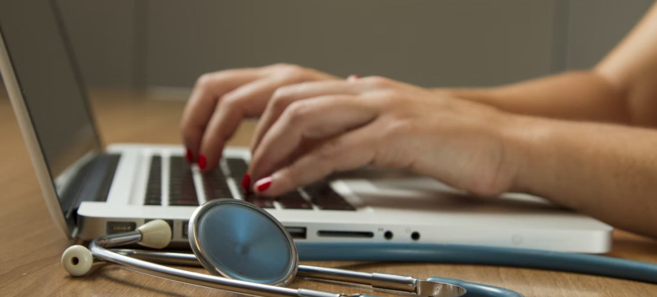 A healthcare administrator works on their laptop with a stethoscope beside them.