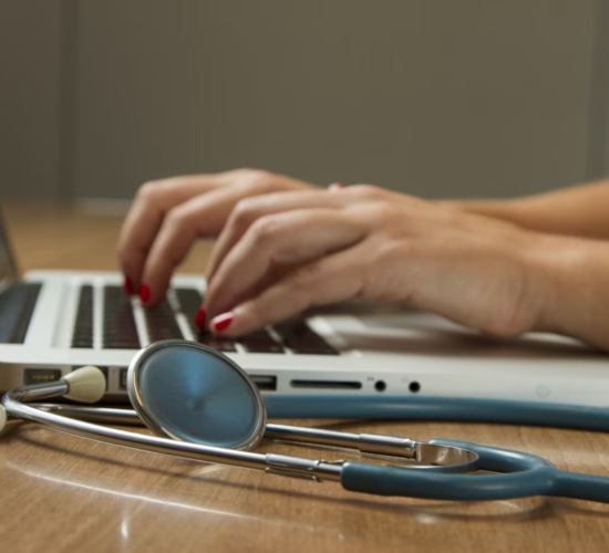 A healthcare administrator works on their laptop with a stethoscope beside them.