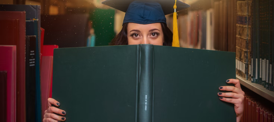 A grad student poses with a large book while wearing a cap.