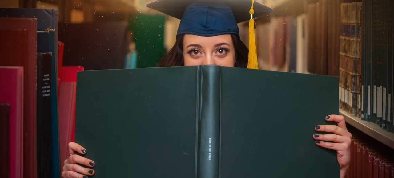 A grad student poses with a large book while wearing a cap.