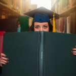A grad student poses with a large book while wearing a cap.