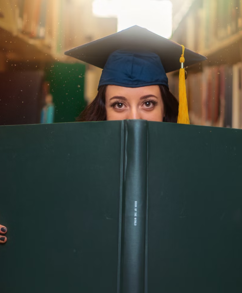 A grad student poses with a large book while wearing a cap.