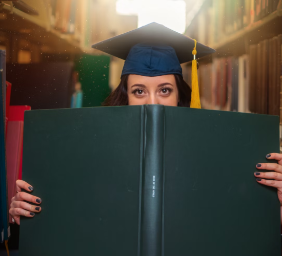 A grad student poses with a large book while wearing a cap.