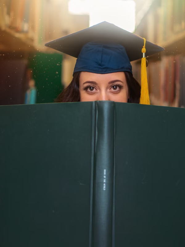 A grad student poses with a large book while wearing a cap.