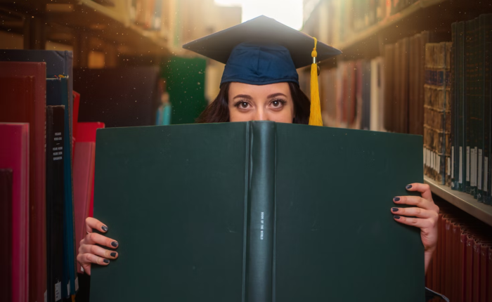 A grad student poses with a large book while wearing a cap.