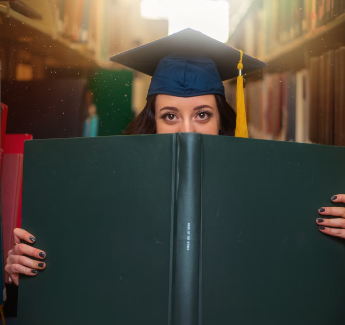 A grad student poses with a large book while wearing a cap.