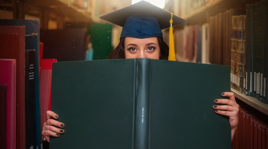 A grad student poses with a large book while wearing a cap.